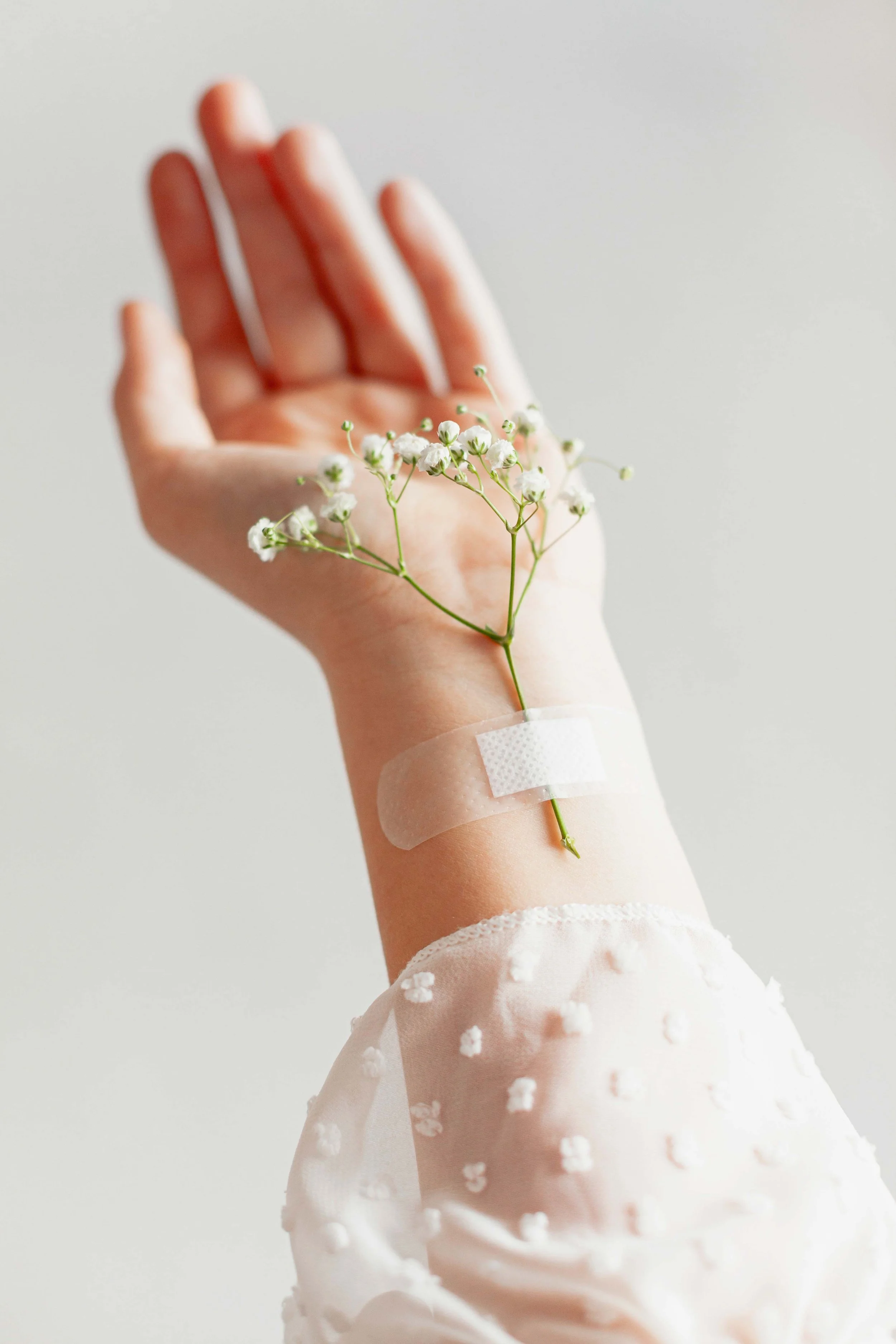 A hand with a hospital IV line and a small bouquet of white baby's breath flowers taped on the wrist, against a plain background.