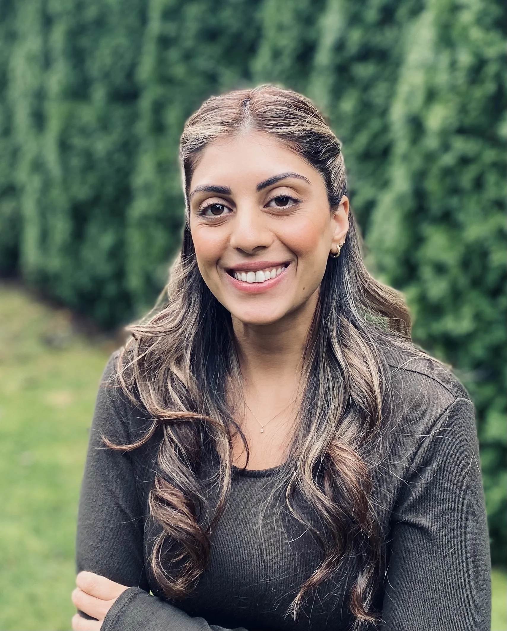 A smiling young woman with long wavy hair standing outdoors with a blurred green hedge background.