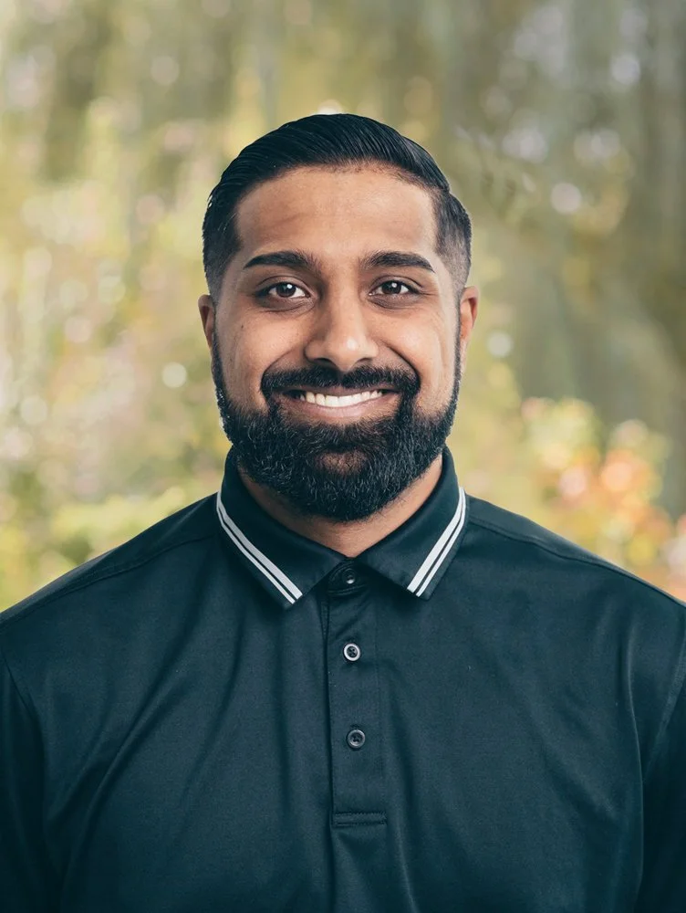 A man with dark hair, a beard, and a mustache smiling outside, wearing a black collared shirt with white stripes on the collar, blurred trees and sunlight in the background.