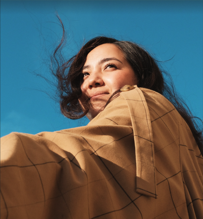 A young woman outdoors with a blue sky background, smiling and looking up, wearing a tan plaid coat.