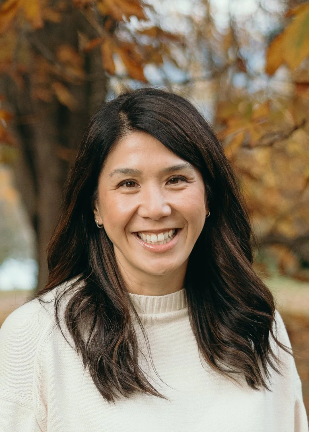 A woman with long dark hair smiling outdoors in front of a tree with orange autumn leaves.