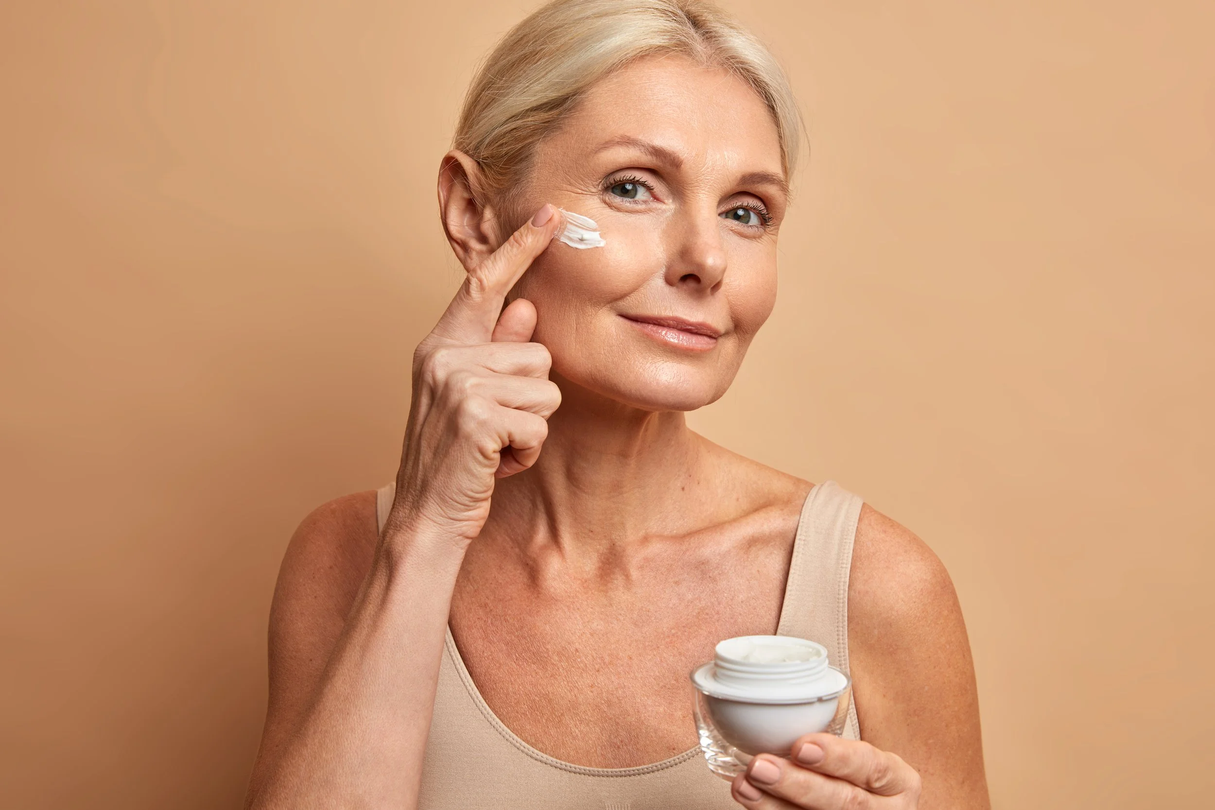 An older woman with short blonde hair applying moisturizer to her face from a small glass bowl, smiling softly against a neutral beige background.