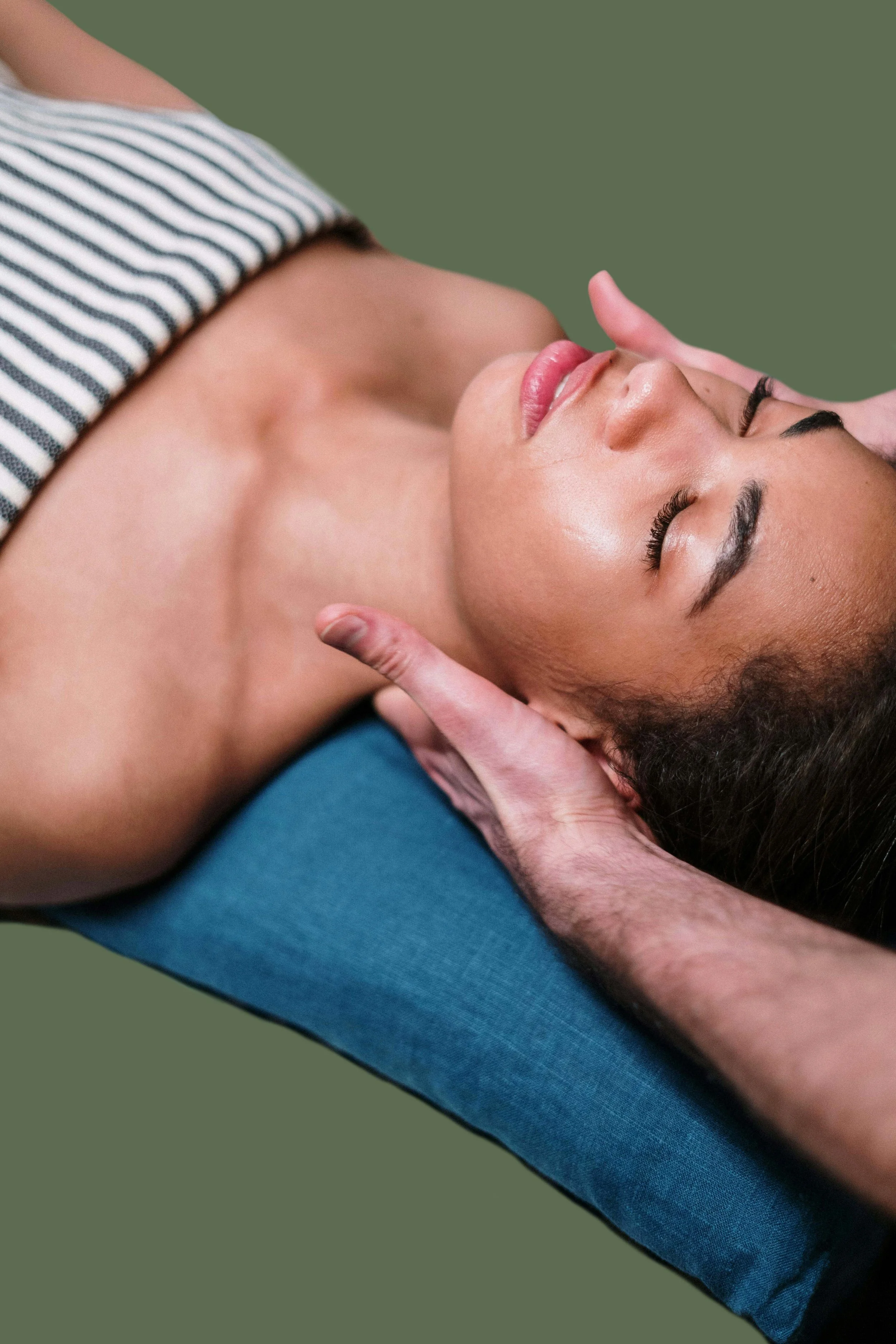 A woman receiving a facial massage from a massage therapist, lying on a blue massage table with a green background.