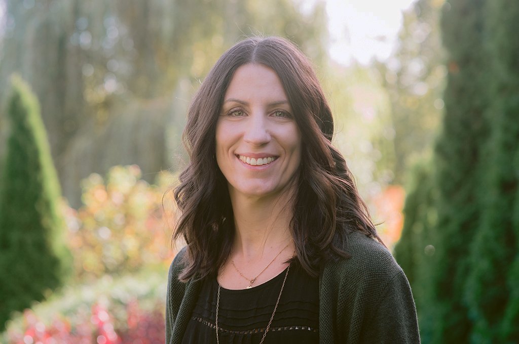 A woman with long dark hair smiling outdoors with blurred trees and greenery in the background.