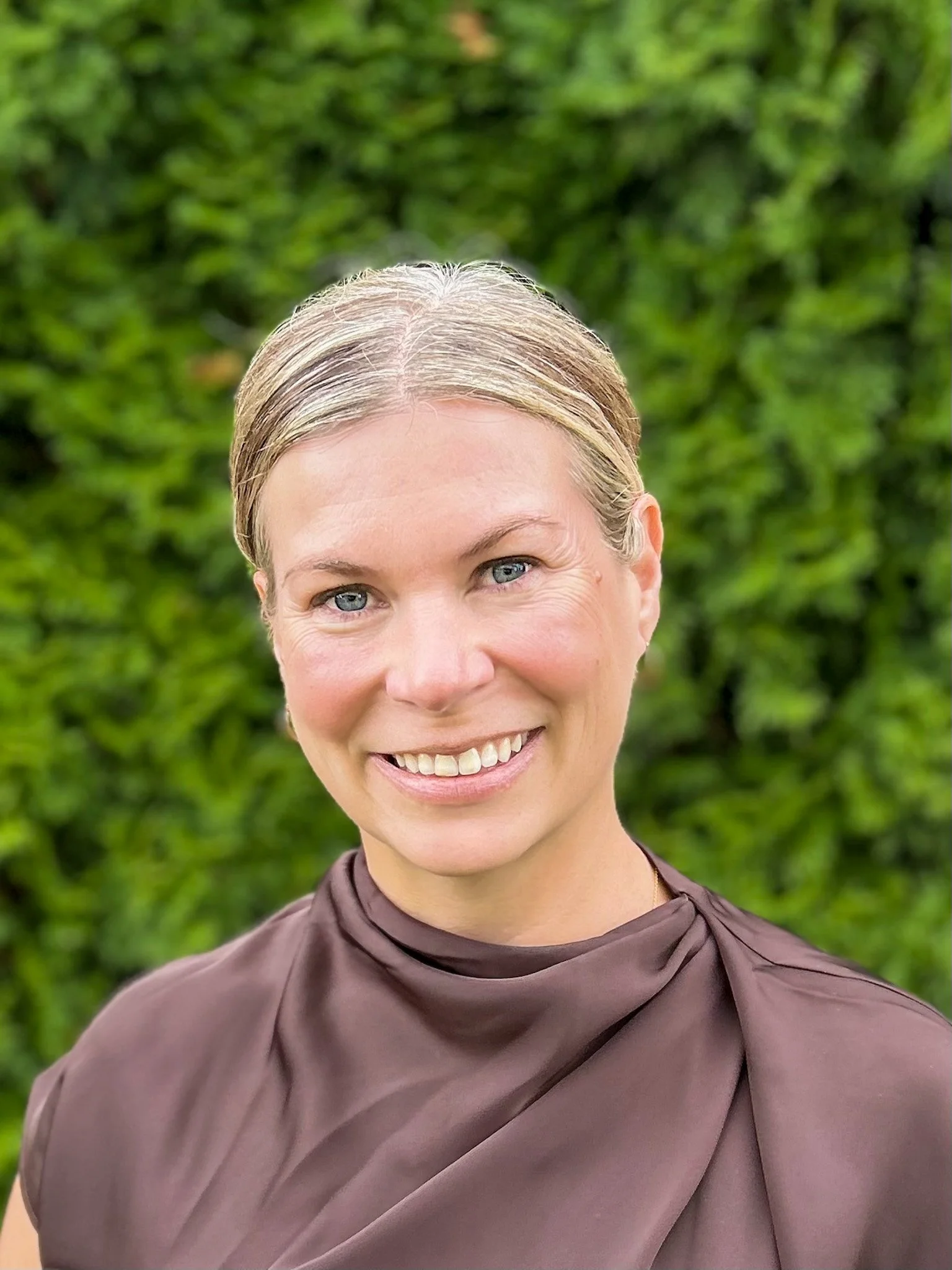 Raeghan Siemens, RAc., Doula, standing outdoors with a green leafy background.