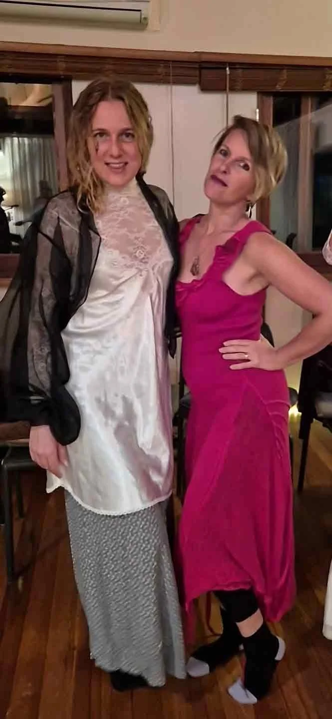 Two women standing together indoors, one wearing a white satin dress with a lace top and patterned skirt, the other in a pink dress with ruffled straps, black socks, and sneakers, posing for the camera.