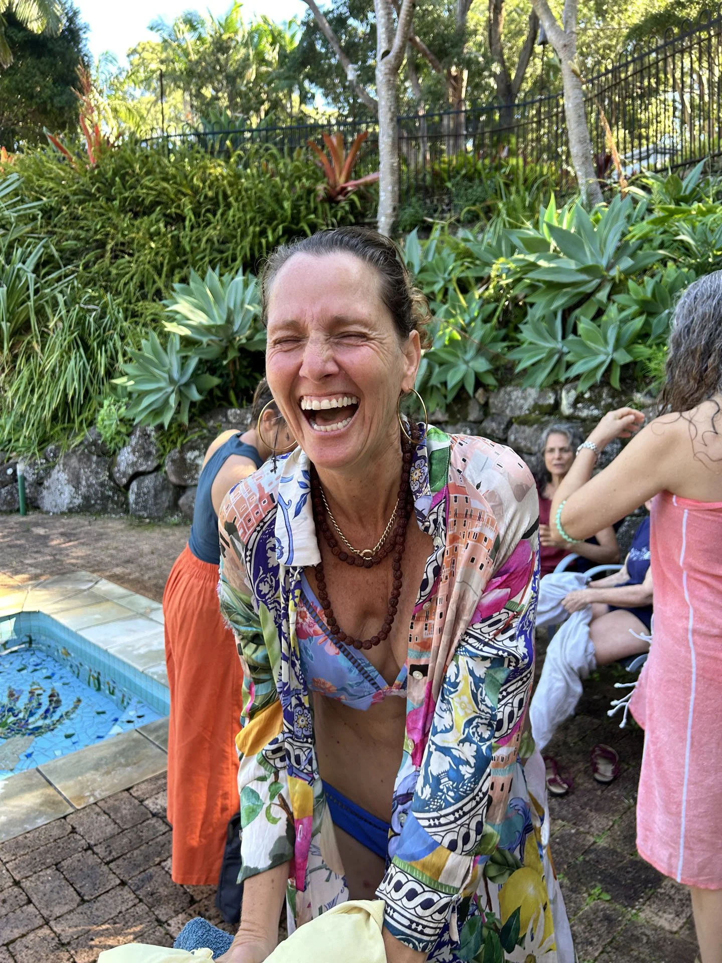 A woman with a big smile, wearing colorful clothing and jewelry, enjoying an outdoor gathering with greenery and other people in the background.