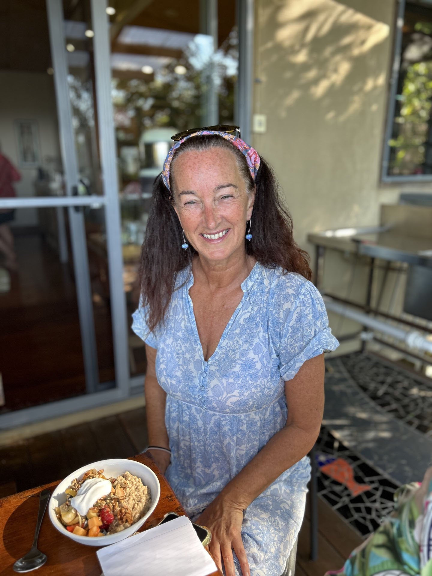 A smiling woman with long dark hair, wearing a blue floral dress, sitting at a table with a bowl of oatmeal topped with fruit and yogurt, in an outdoor setting with shaded and sunlit areas.