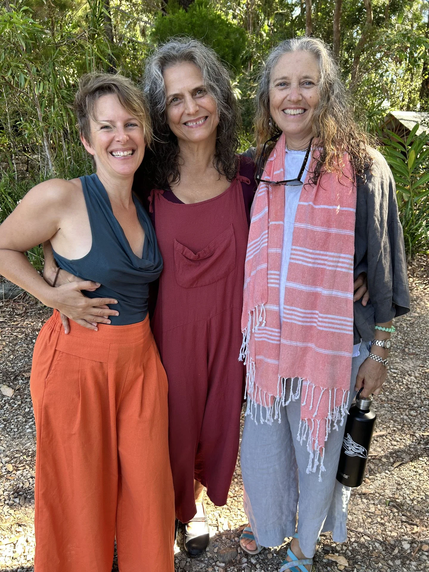 Three women standing outdoors amid trees, smiling at the camera. The woman on the left wears an orange skirt and a sleeveless top, the woman in the middle wears a red dress, and the woman on the right wears a gray skirt with a pink scarf, glasses aro