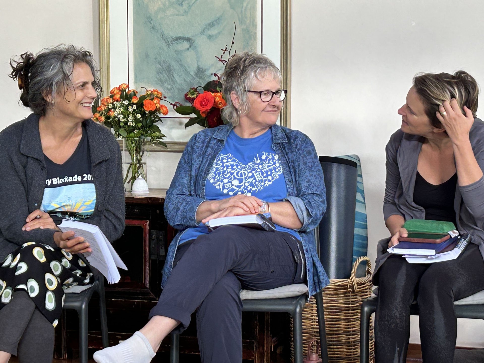 Three women are engaged in conversation in a room with a bouquet of orange and white flowers on a table behind them. The woman in the middle is wearing a blue shirt with musical notes and glasses. The woman on the right is touching her hair and holding a tablet. The woman on the left is smiling and holding papers.