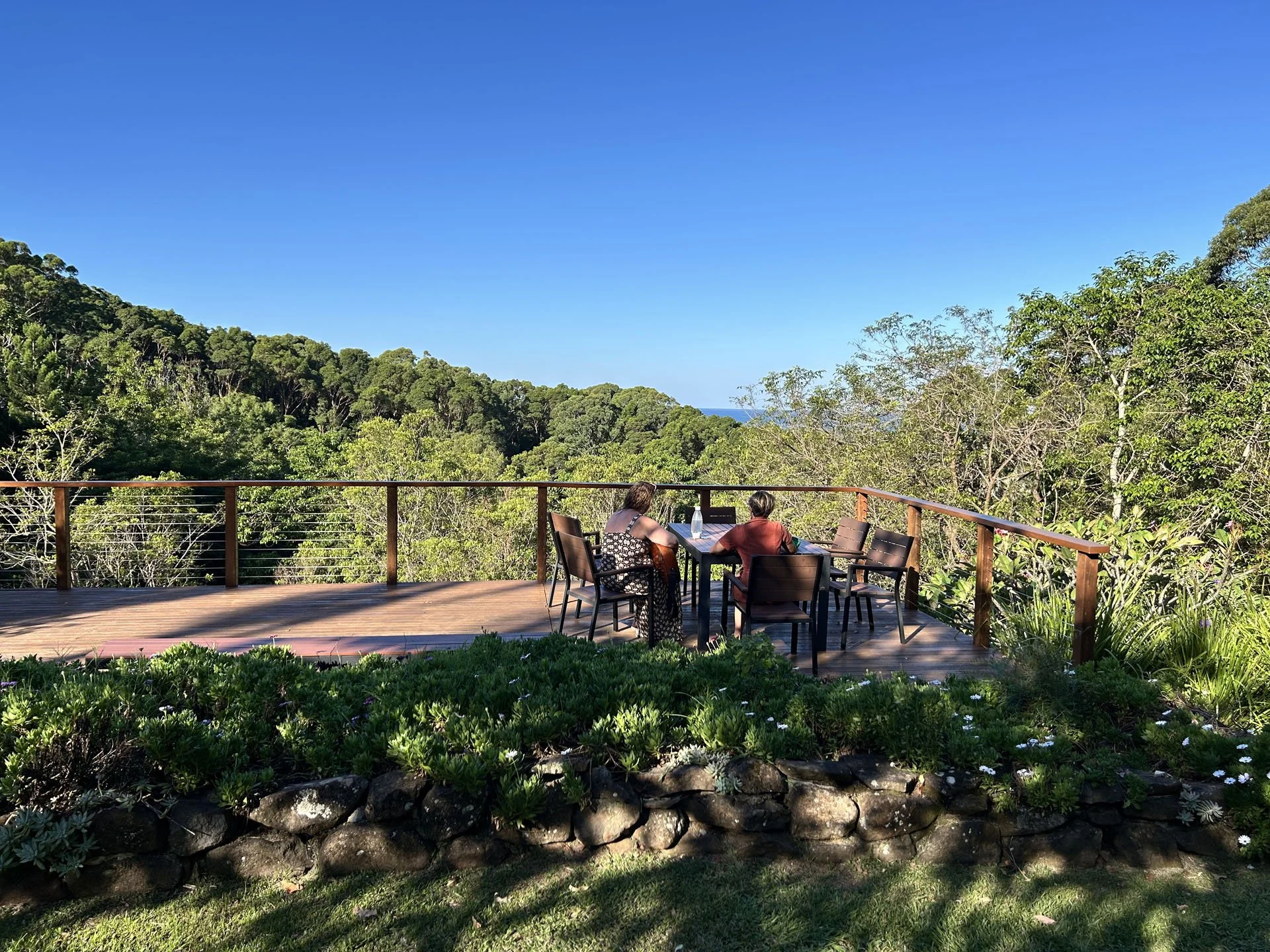 People sitting at a table on a wooden deck overlooking a lush green forest with trees, hills, and a clear blue sky in the background.