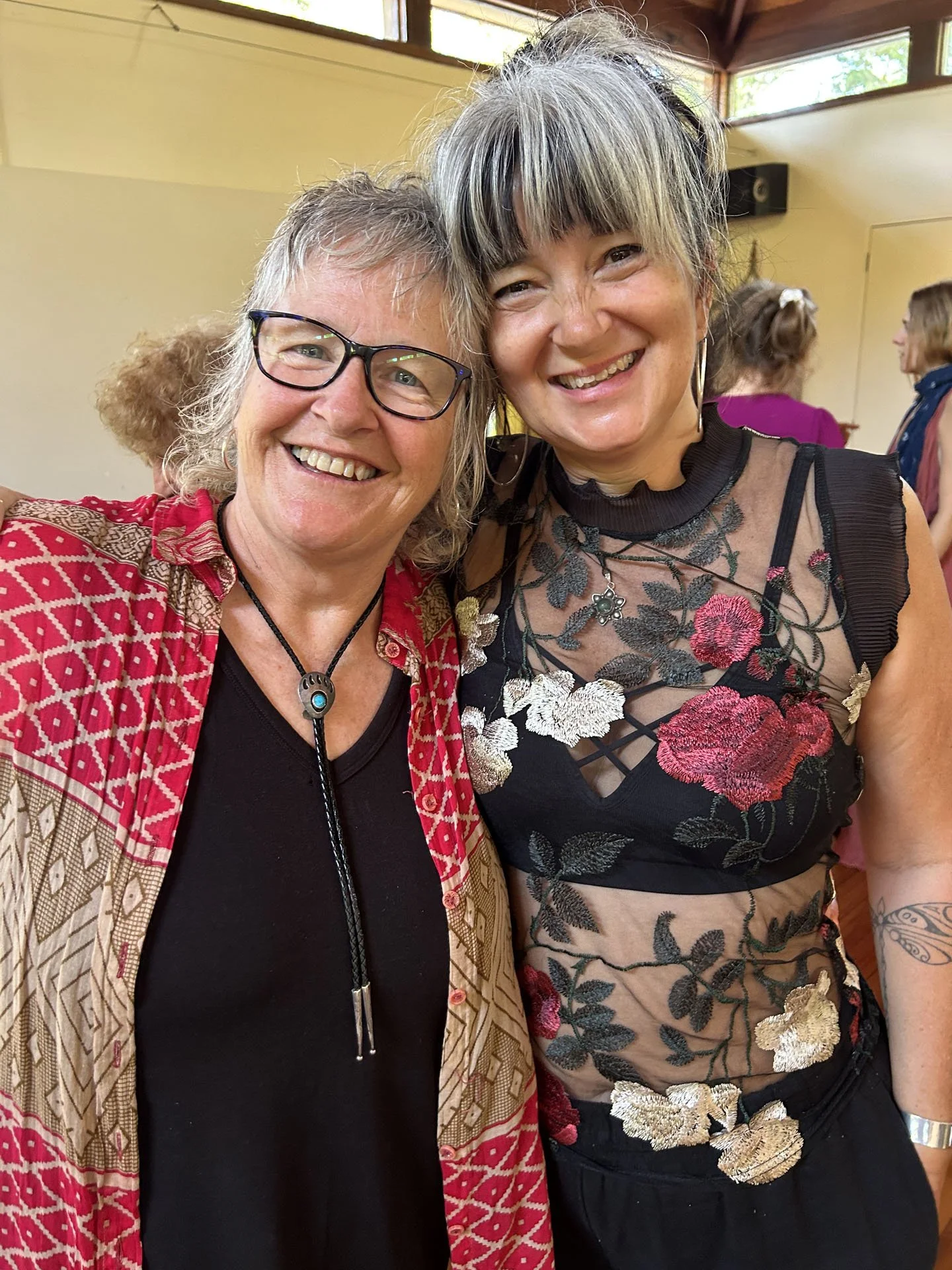 Two women smiling closely at the camera, one wearing glasses and a patterned red jacket, and the other wearing a floral sheer top, in a warmly lit room with other people in the background.