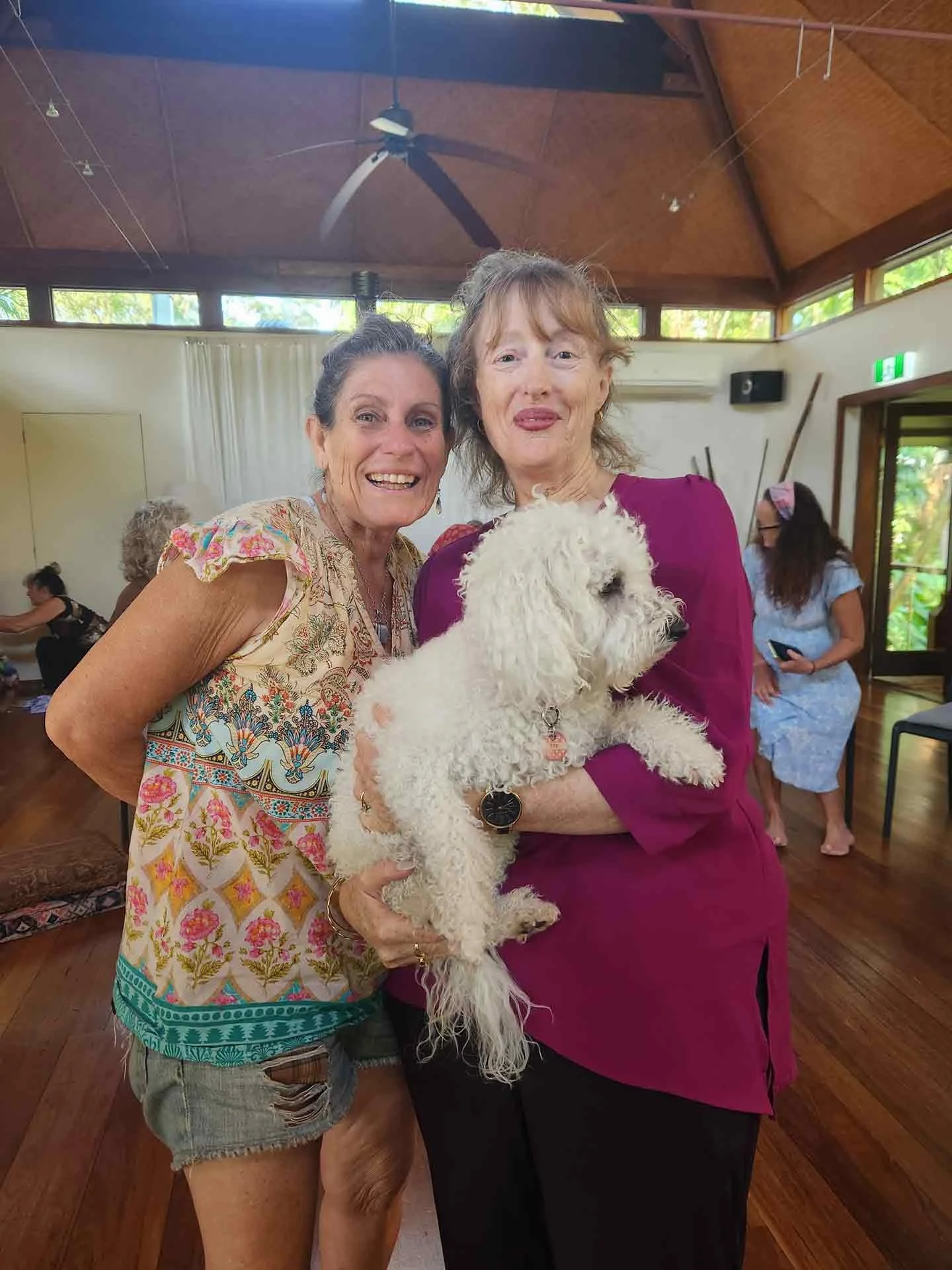 Two women smiling and holding a white fluffy dog indoors, with a woman in the background using a phone, and another woman sitting nearby.