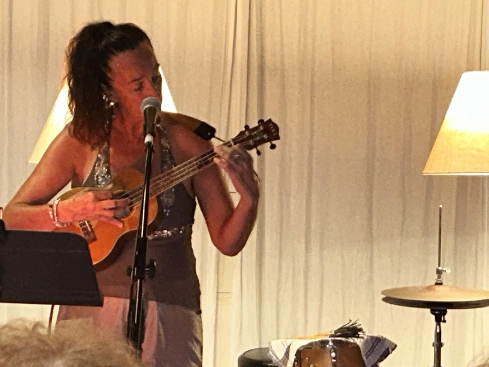 A woman with short dark hair singing into a microphone and playing a ukulele in a room with beige curtains and warm lighting.