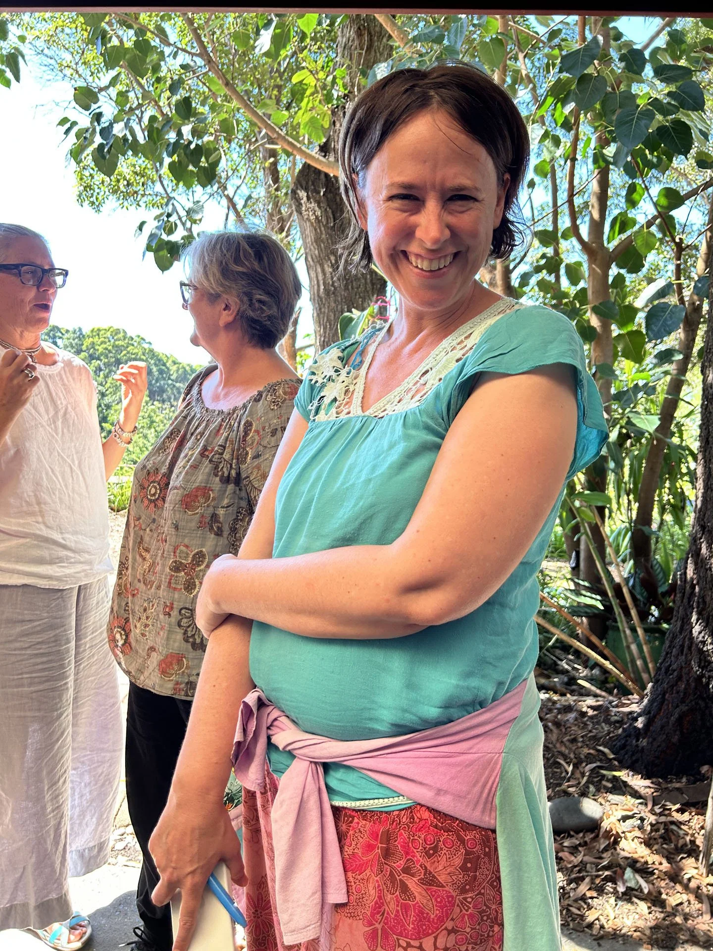 Smiling woman with short dark hair, wearing a turquoise top with lace details, standing outdoors among trees with three other women in the background, engaging in conversation on a sunny day.