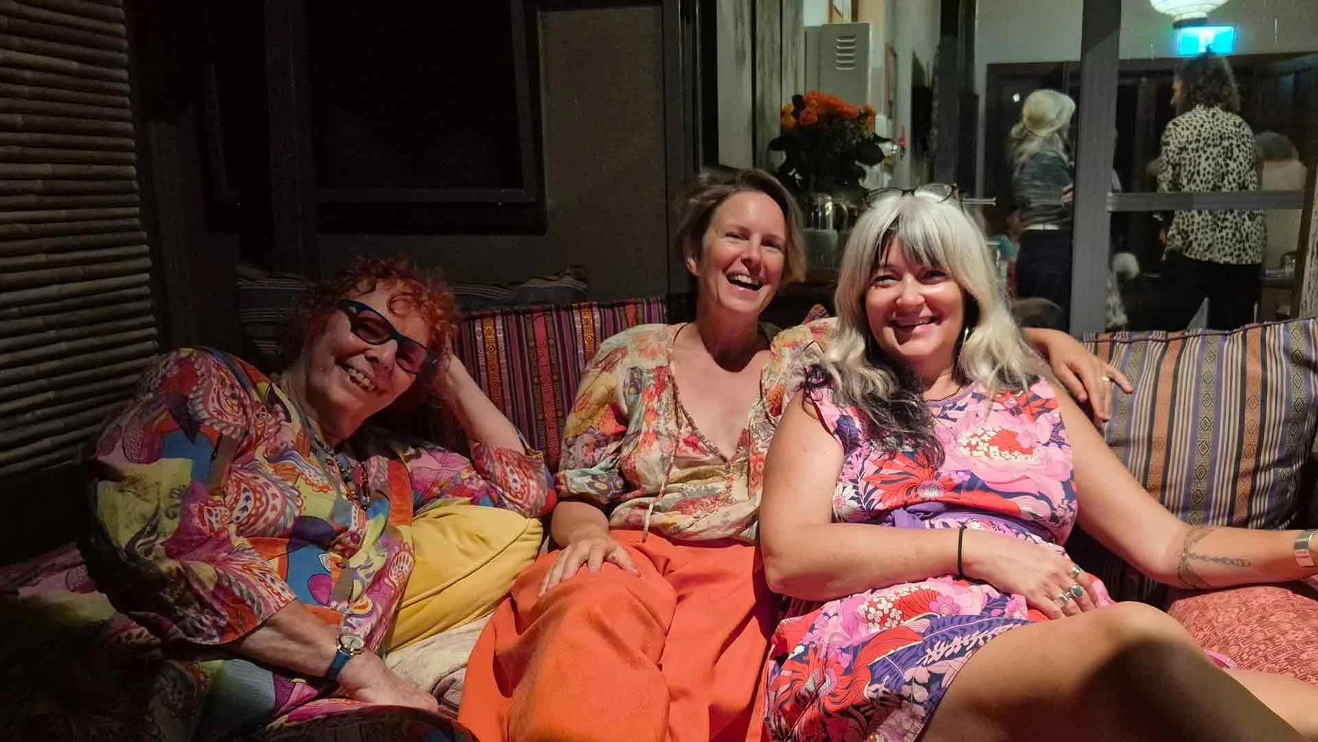 Three women sitting on a colorful striped couch, smiling and enjoying each other's company in an indoor setting at night. They are dressed in vibrant, patterned clothing and appear happy.