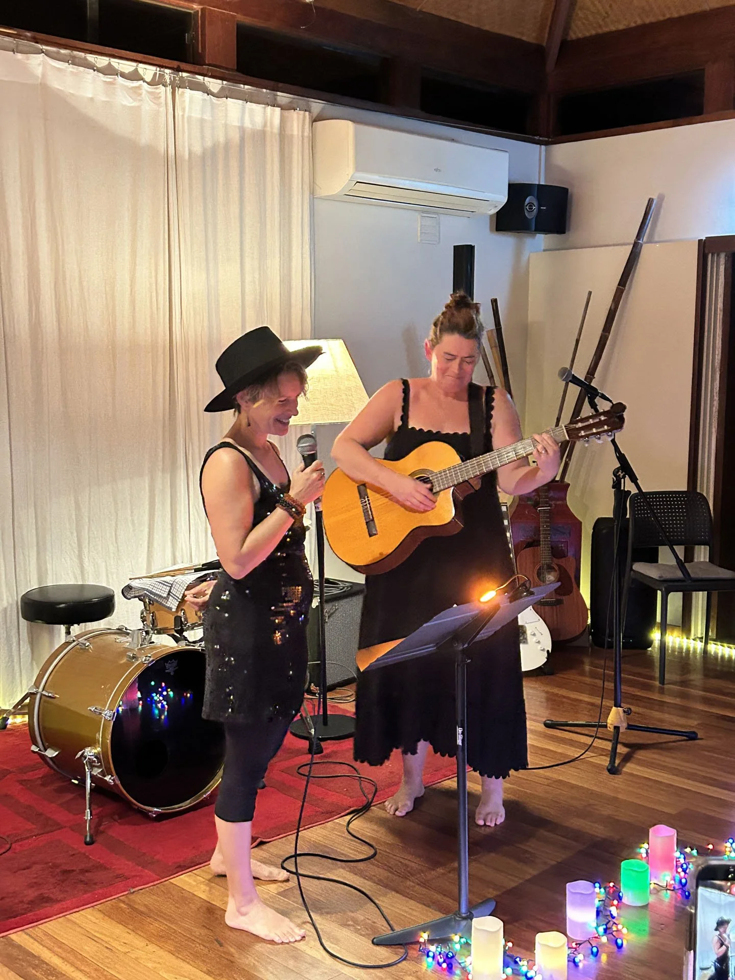 Two women are performing musical acts in a cozy indoor space. One woman is singing into a microphone while the other is playing an acoustic guitar. They are barefoot, and there's a drum kit and a guitar in the background. The scene is decorated with 