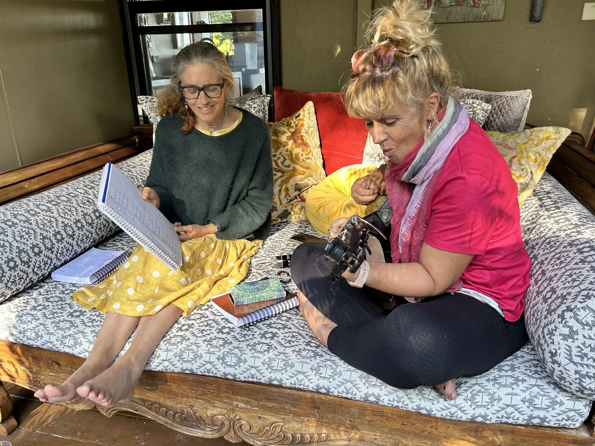 Two women sitting on a wooden and cushioned bench. One woman is holding a notebook, the other woman is looking at a camera. There are various notebooks, papers, and a yellow piece of clothing on the bench. Behind them are decorative pillows and windows with a view of outdoor trees.