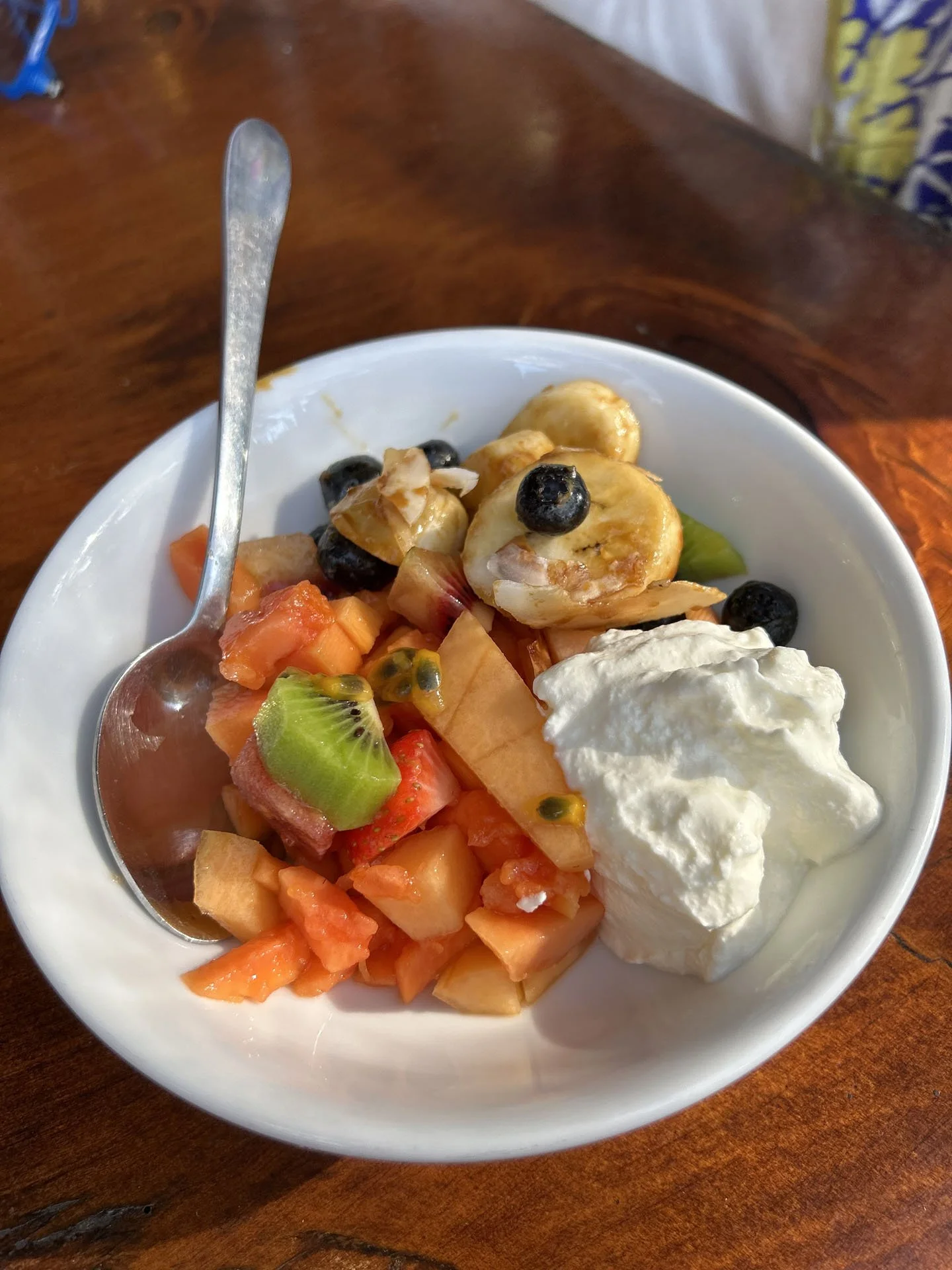 A white bowl filled with various fruits, yoghurt, and a spoon on a wooden table.