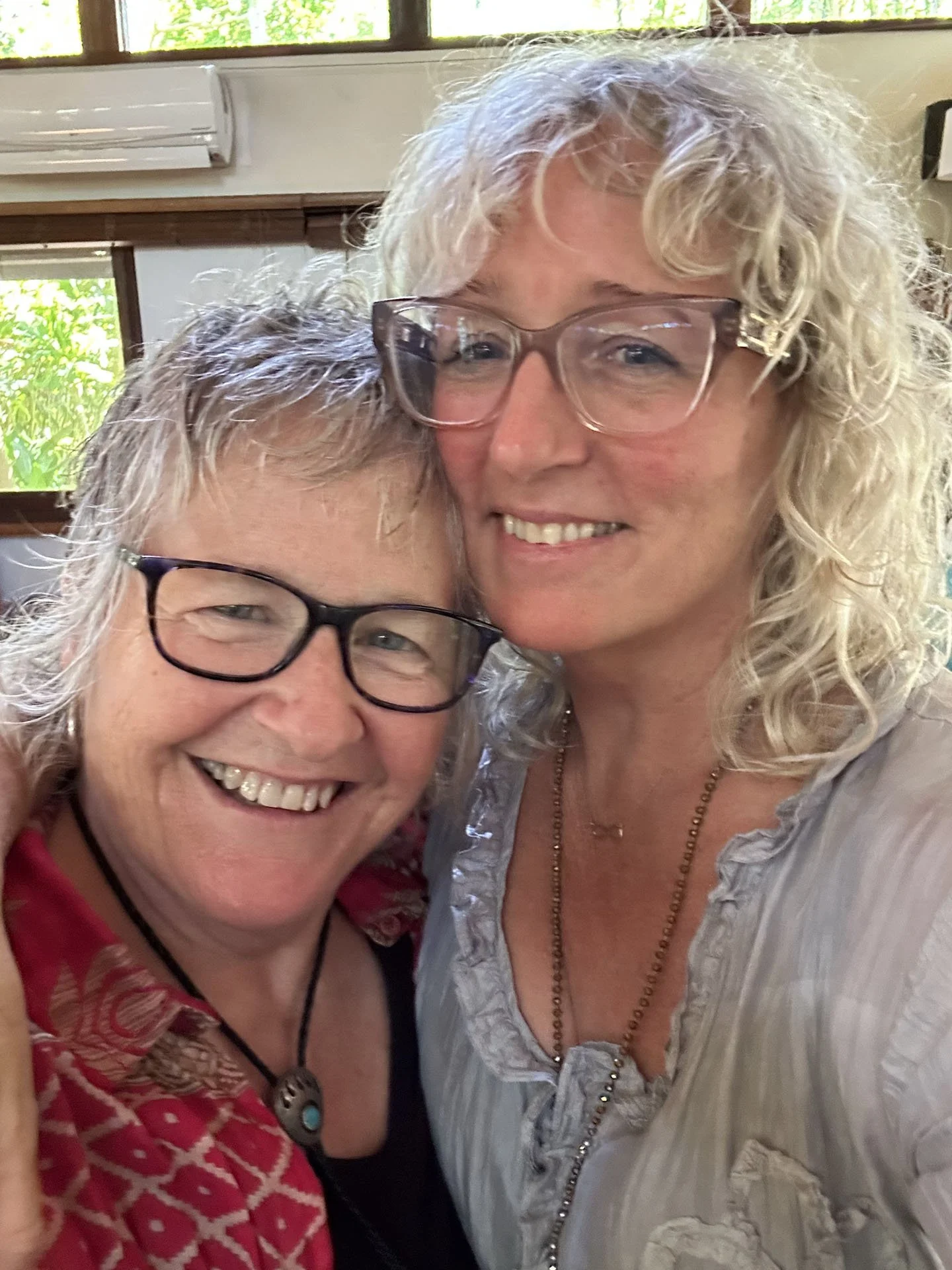 Two women smiling close together indoors with windows and greenery in the background.