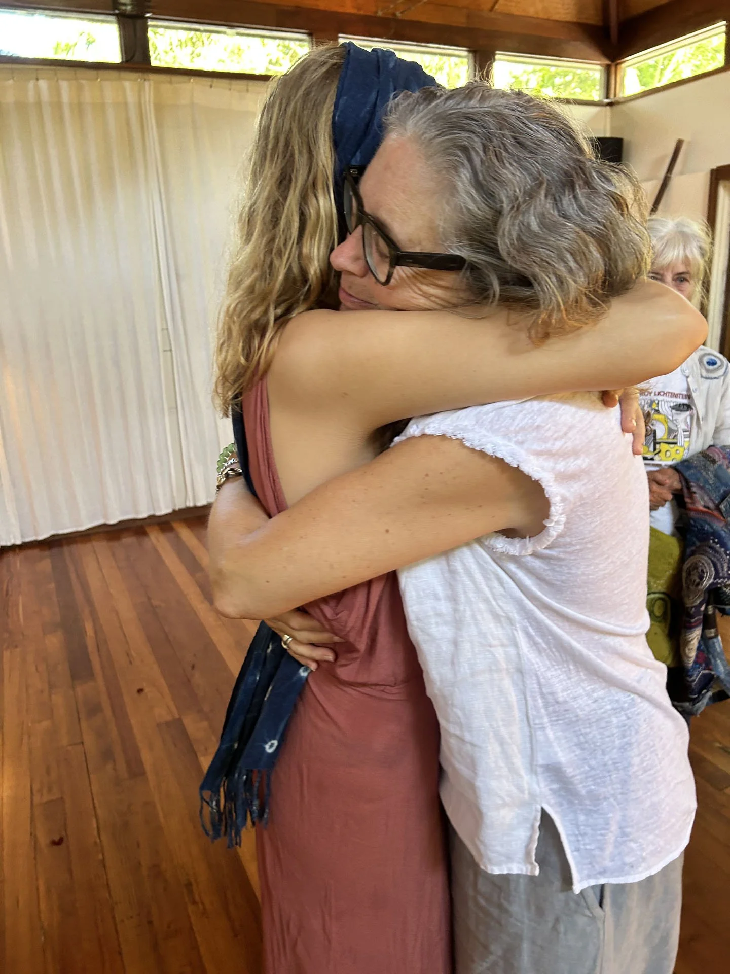 Two women hugging each other warmly inside a room with wooden floors and a large window at the back. One woman has curly blonde hair and glasses, while the other has long blonde hair and wears a reddish-brown dress.