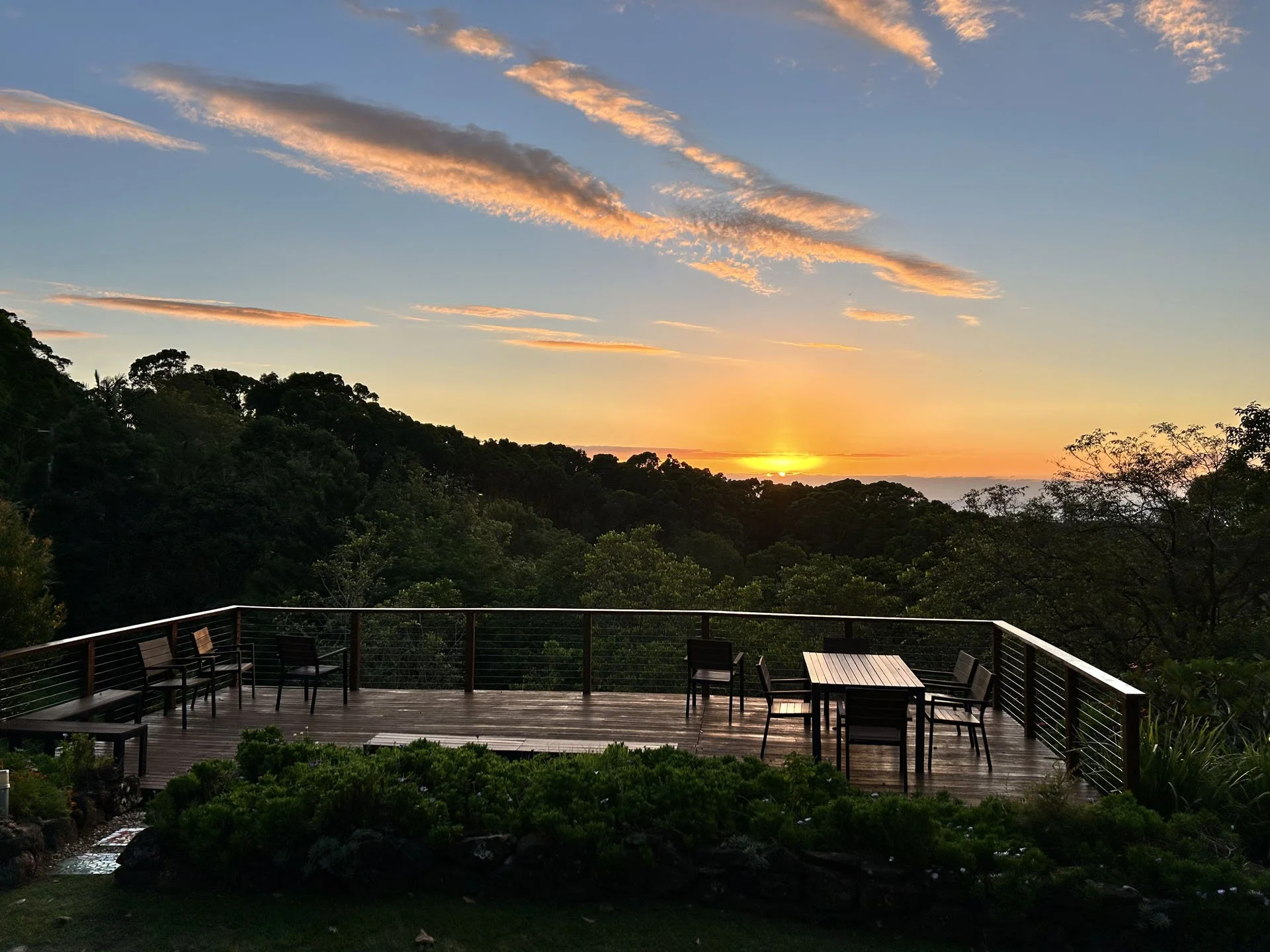 A deck with chairs and a table overlooking a forested area at sunset, with clouds and the sun near the horizon.