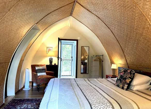 Bedroom with a curved ceiling, bed with patterned bedding, side table with lamp, armchair, floor lamp, door to balcony, mirror, and small wardrobe.