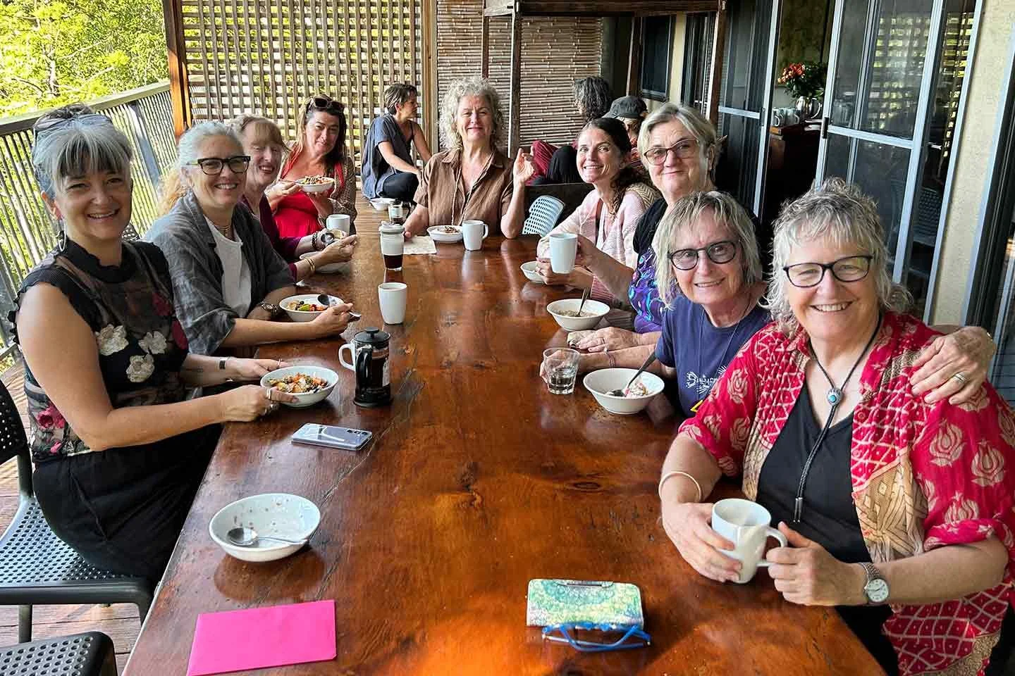 Group of women sitting around a large wooden table on a balcony, enjoying breakfast together with bowls of food, coffee, and drinks, smiling at the camera.