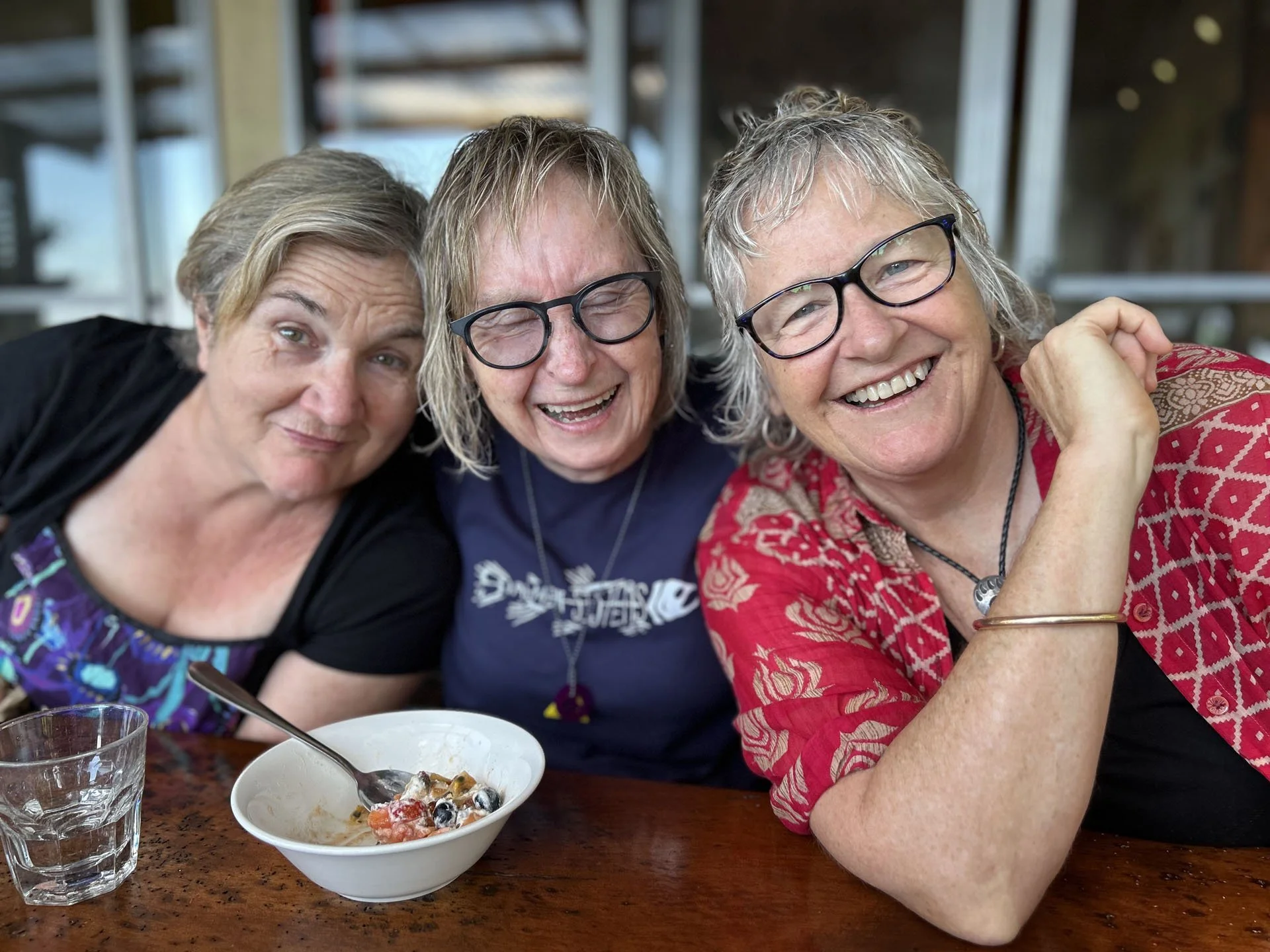 Three women sitting at a table, smiling and laughing together. A bowl of fruit and a glass of water are in front of them.