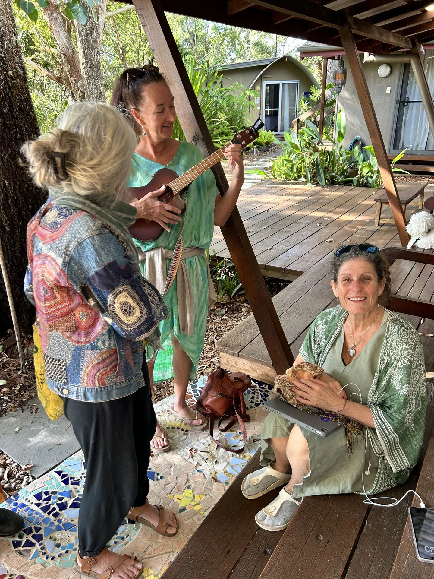 Three women in an outdoor setting, with one playing a ukulele, another sitting at wooden stairs with a small dog, and a third woman looking on. There is a colourful tiled floor, plants, and a wooden deck area in the background.