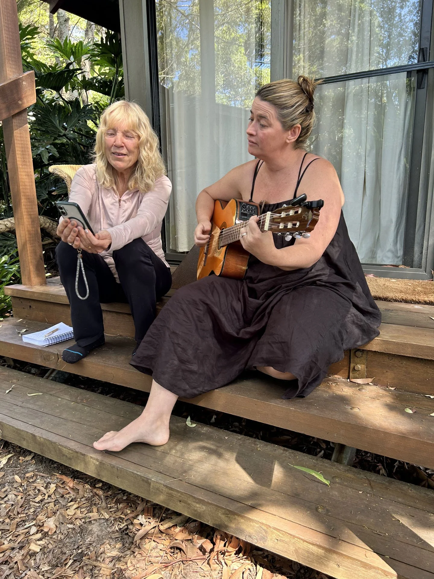Two women sitting on a wooden porch outdoors, one playing guitar and the other holding a phone, with one woman appearing emotional and the other looking contemplative.