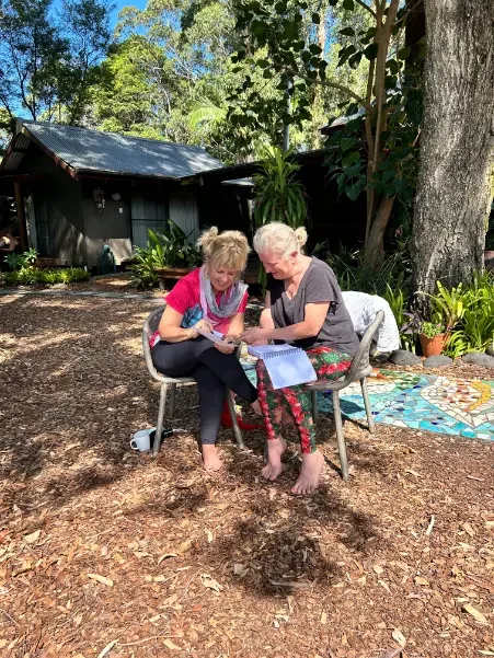 Two elderly women sitting outdoors, looking at a tablet together in a garden with trees and plants, a shed in the background, and a colorful mosaic on the ground.