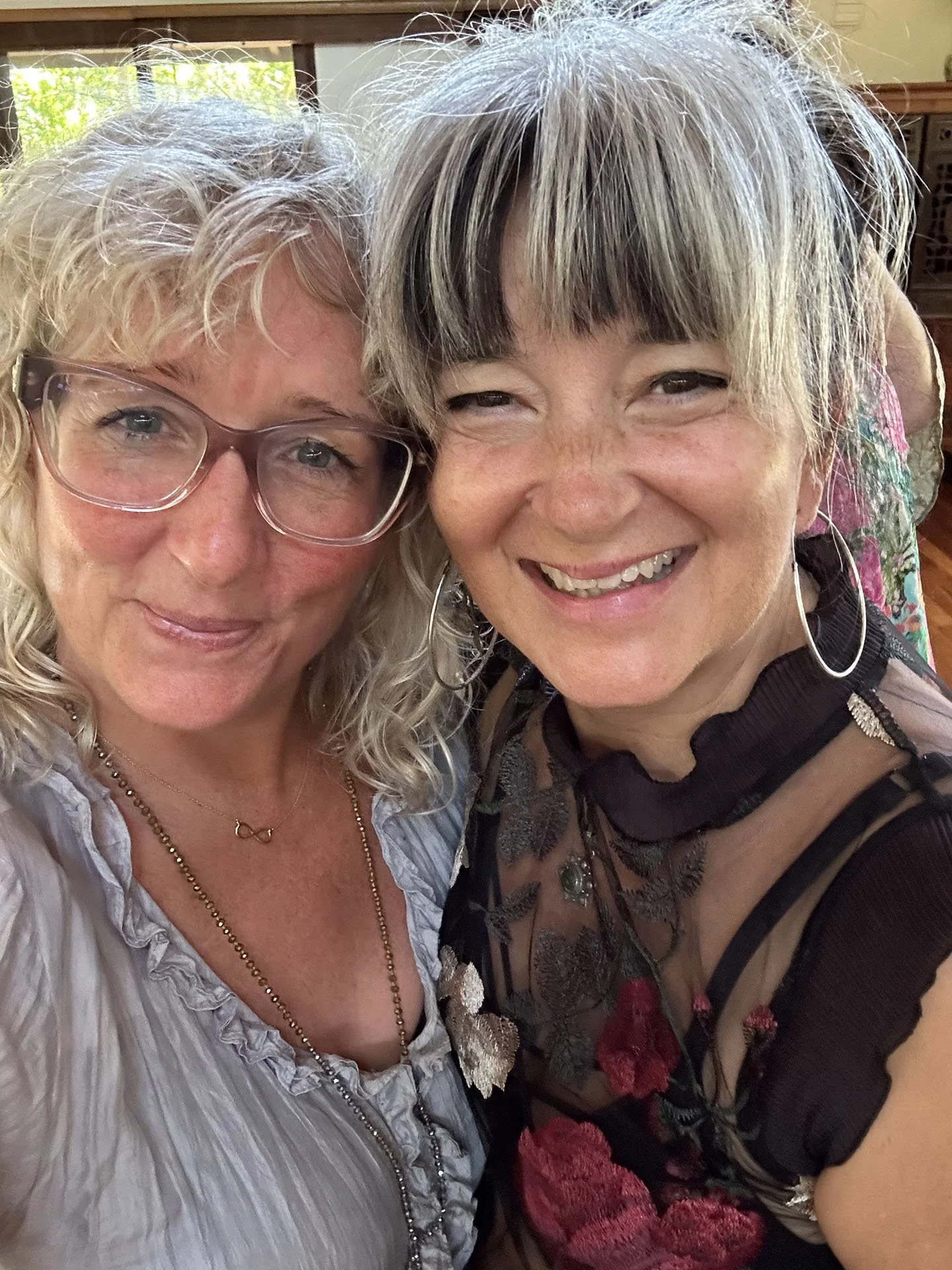 Two women smiling closely together, one wearing glasses and a light blouse with a necklace, the other with short hair, earrings, and a black dress with red accents, in a room with a window and wooden furniture.