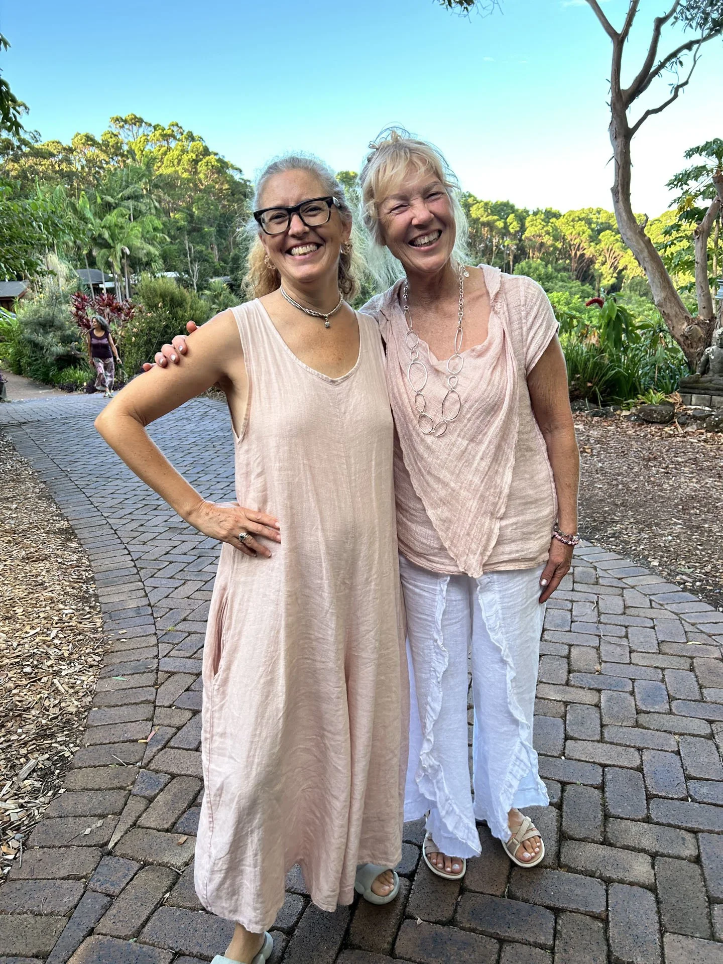Two women standing outdoors on a paved path, smiling and posing for the photo, with lush greenery and trees in the background.