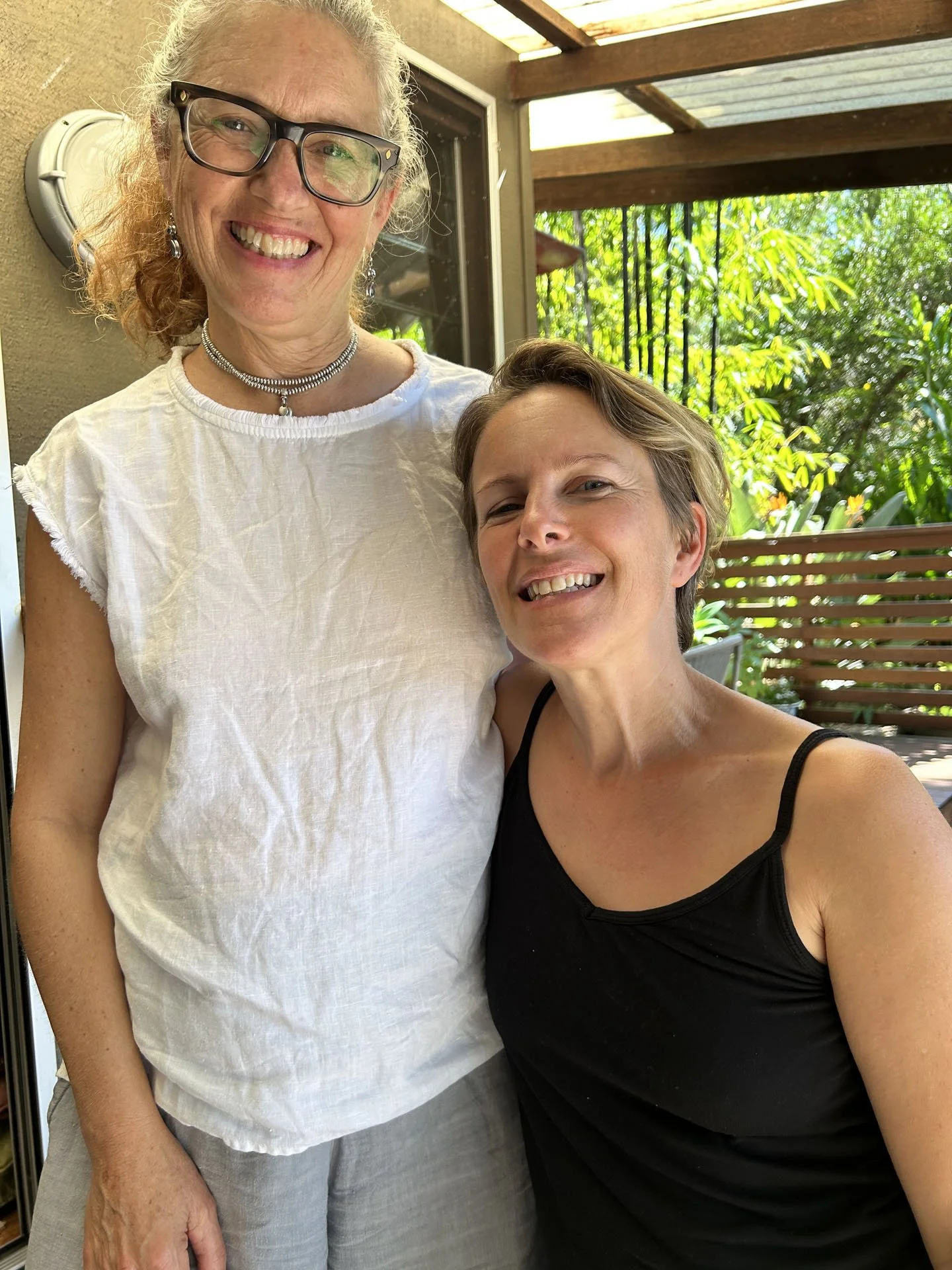 Two women smiling, standing close together on a patio with greenery in the background.