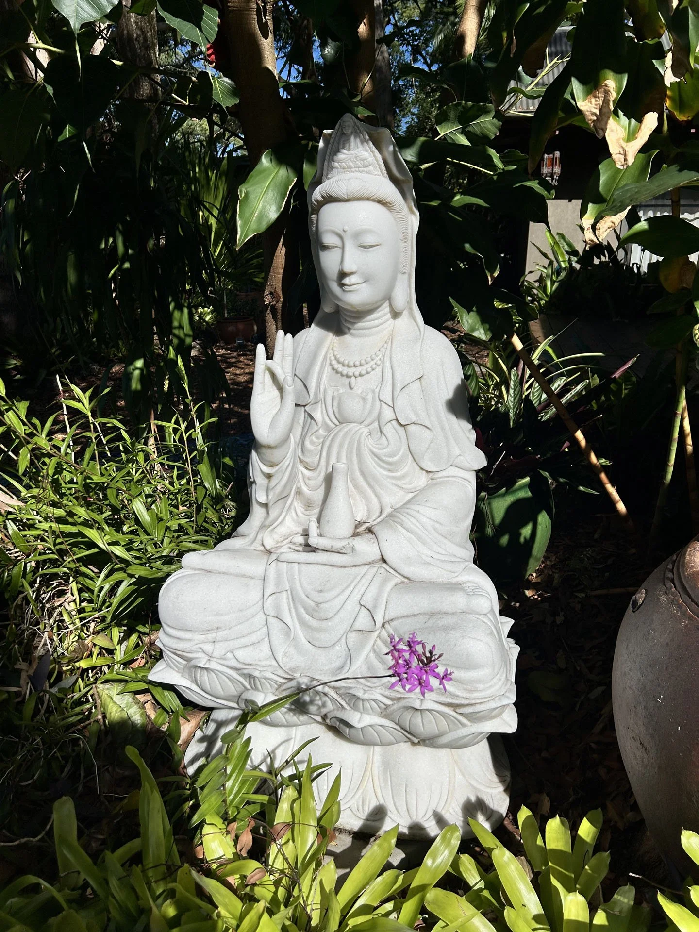 White marble statue of a serene female figure, possibly a bodhisattva, seated outdoors among green plants with purple flowers at her feet, and a backdrop of trees and a building.