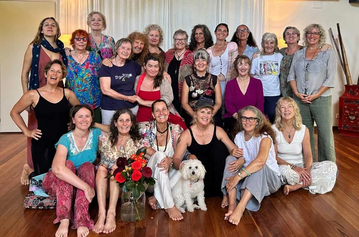 Group of 20 women and one white dog indoors, some sitting on the floor and others standing behind, in a room with wooden floor, beige walls, and curtains.