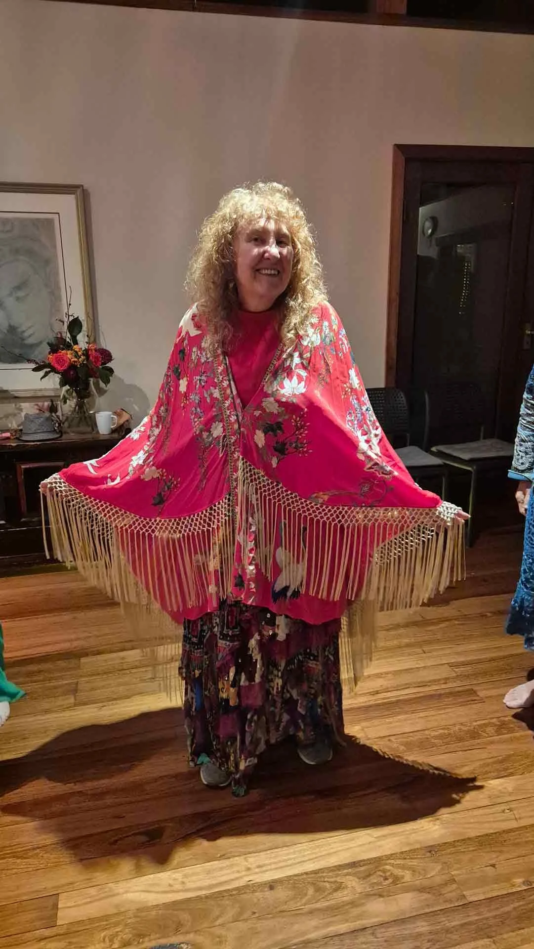 A woman with curly blonde hair smiling and standing indoors, wearing a bright pink shawl with floral patterns and fringe, over a matching pink top, and patterned pants.