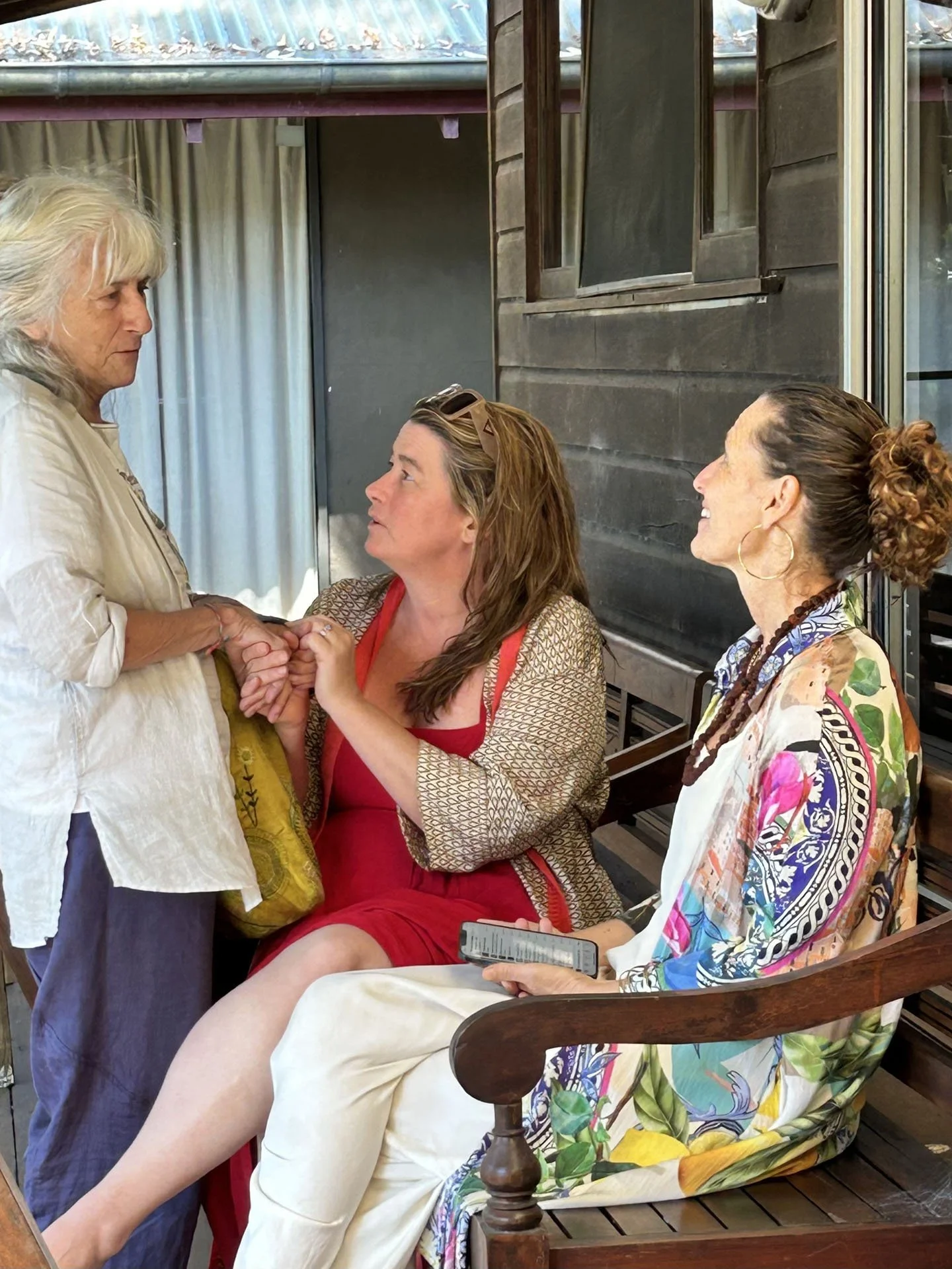 Three women engaged in a conversation on a wooden porch. Two women are seated, one standing, holding the hands of the woman in red. The woman in white and the woman in patterned dress are smiling. One woman is holding a smartphone.