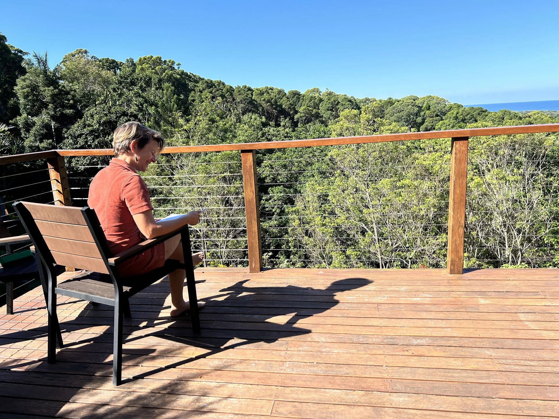A person with short hair sitting on a wooden deck, reading a book, with green trees and a blue sky in the background.