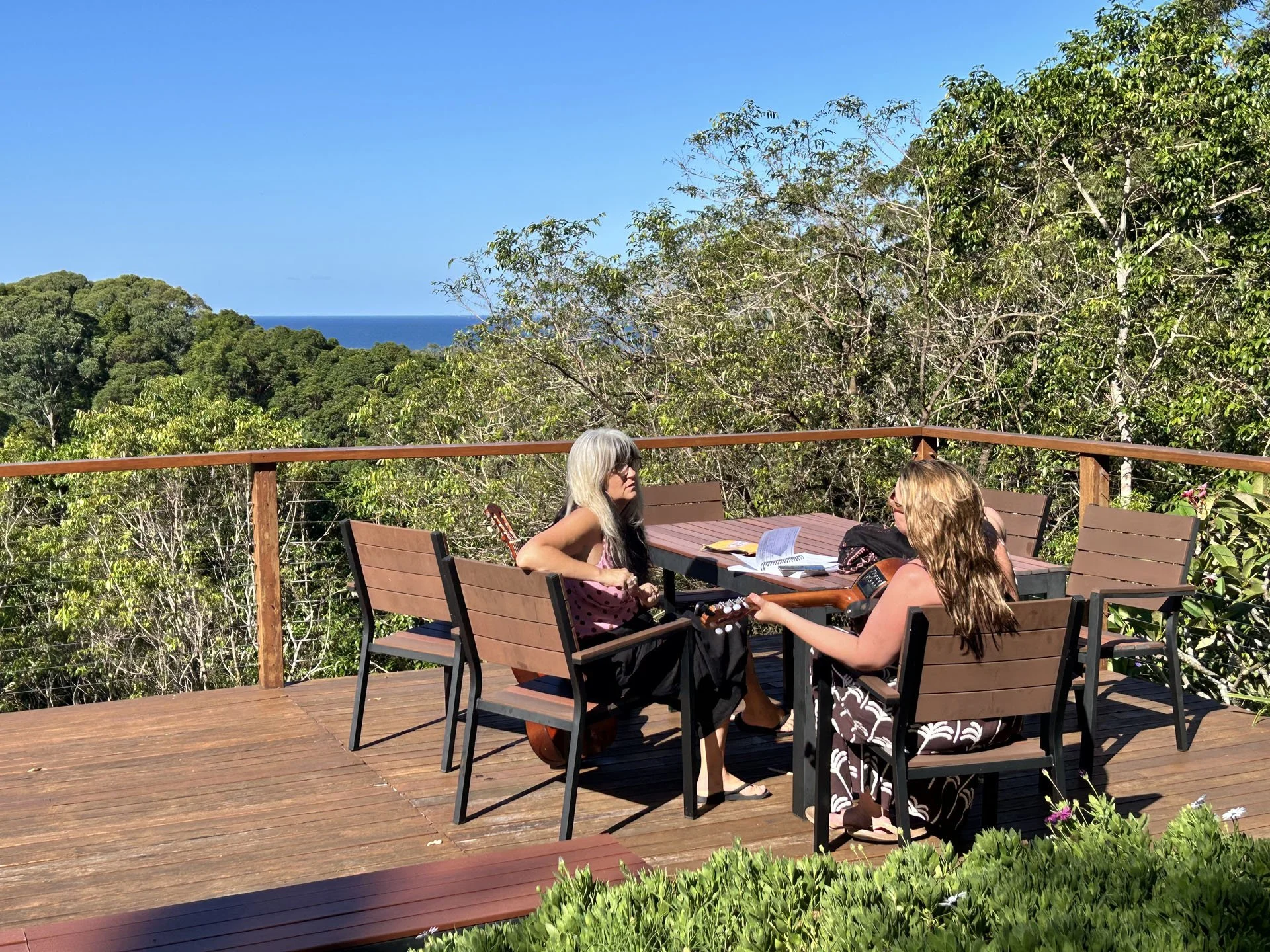 Three women sitting at a wooden outdoor table on a deck surrounded by lush green trees, with the ocean visible in the background on a sunny day.