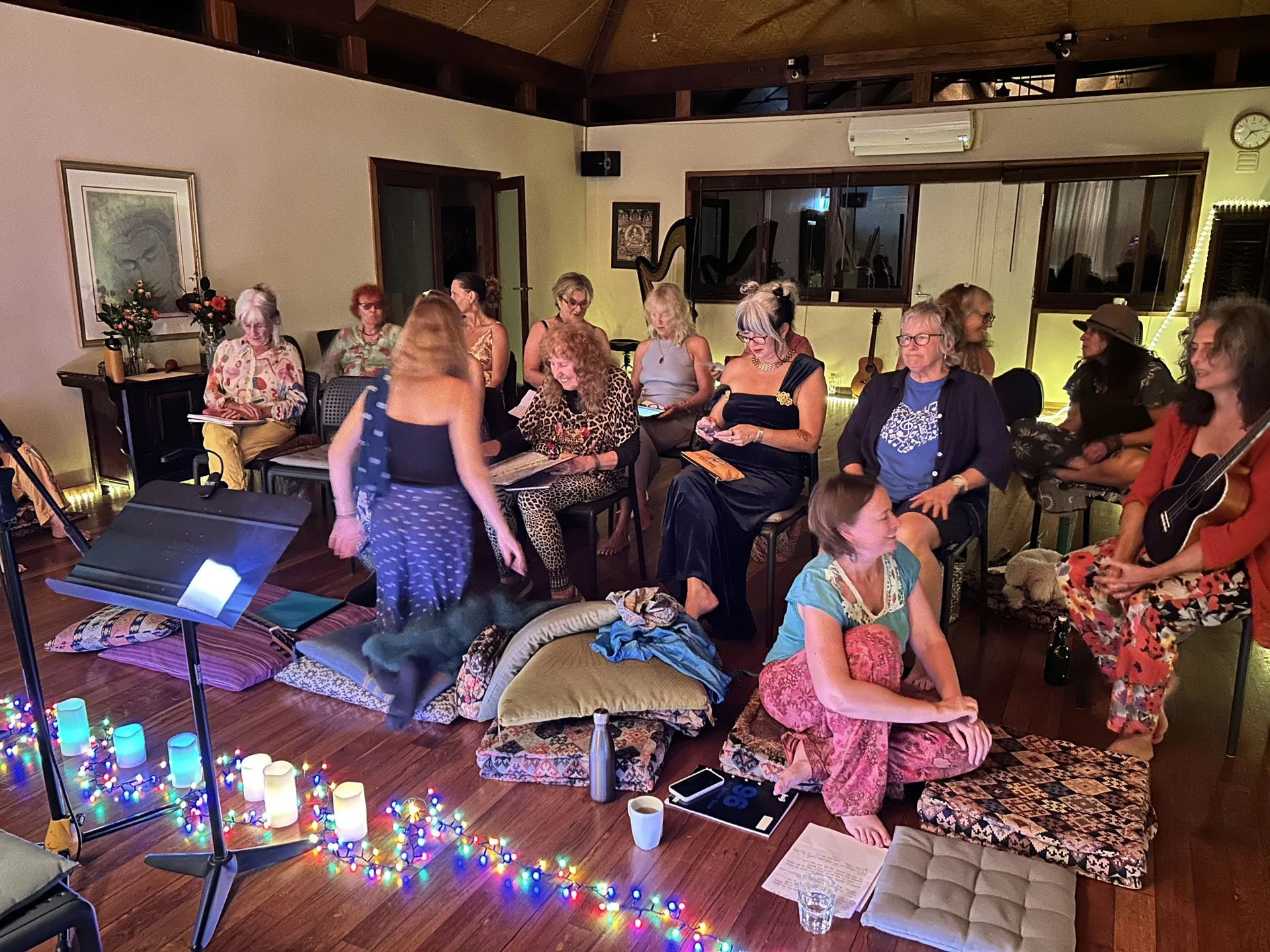 A group of mostly women sitting on chairs and cushions in a cozy indoor setting, with some musical instruments visible, including a guitar. There is a shelf with candles and string lights on the floor, and a music stand in the foreground. The room has warm lighting, decorated with artwork, flowers, and an air conditioning unit.