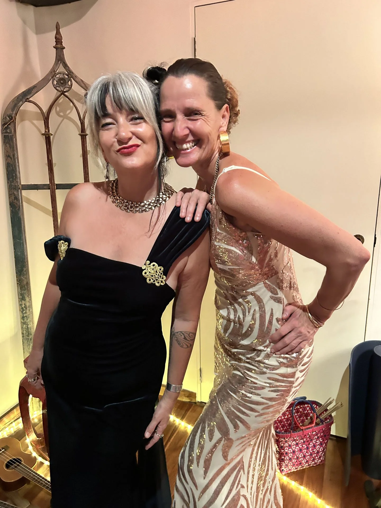 Two women dressed elegantly, smiling and posing for a photo at a celebration or event, with a guitar and string lights in the background.