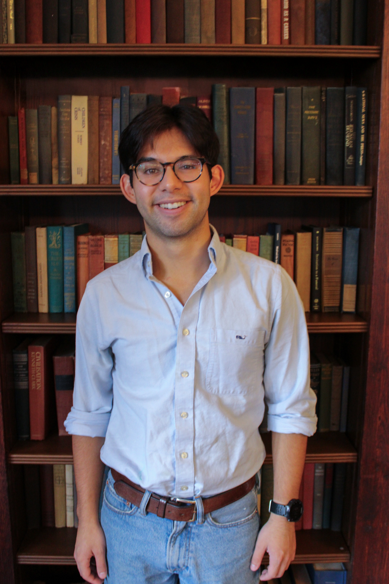 A young man with dark hair, wearing glasses, a light blue button-up shirt, jeans, a brown belt, and a black smartwatch on his left wrist, standing in front of a wooden bookshelf filled with colorful books.