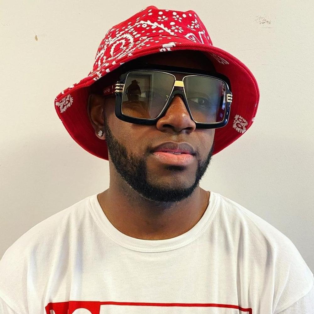 A man wearing a red bucket hat with white patterns, large black sunglasses, a white T-shirt, and earrings, standing against a plain wall.