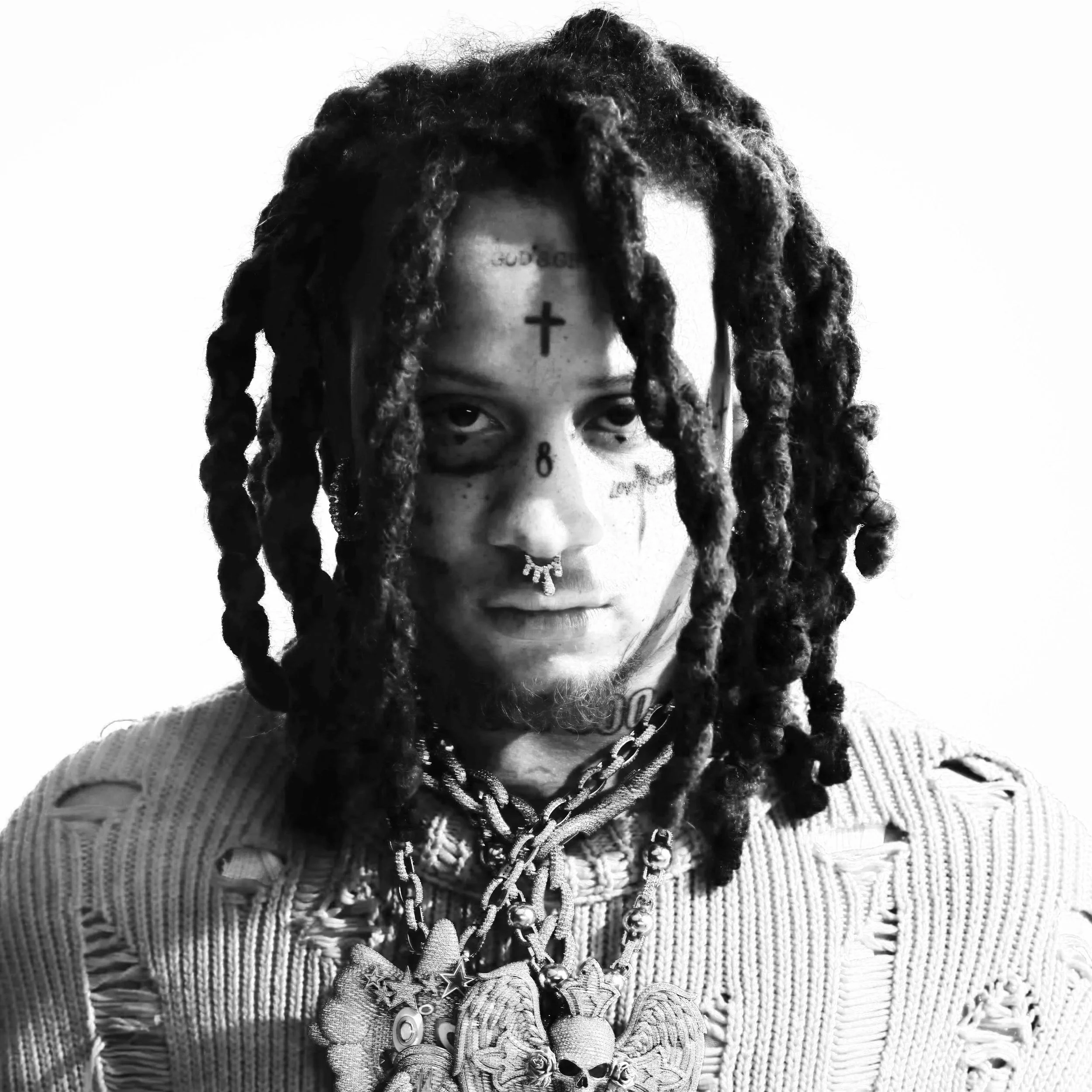 A young man with dreadlocks, tattoos, and multiple necklaces, including a prominent skull pendant, poses against a plain background.