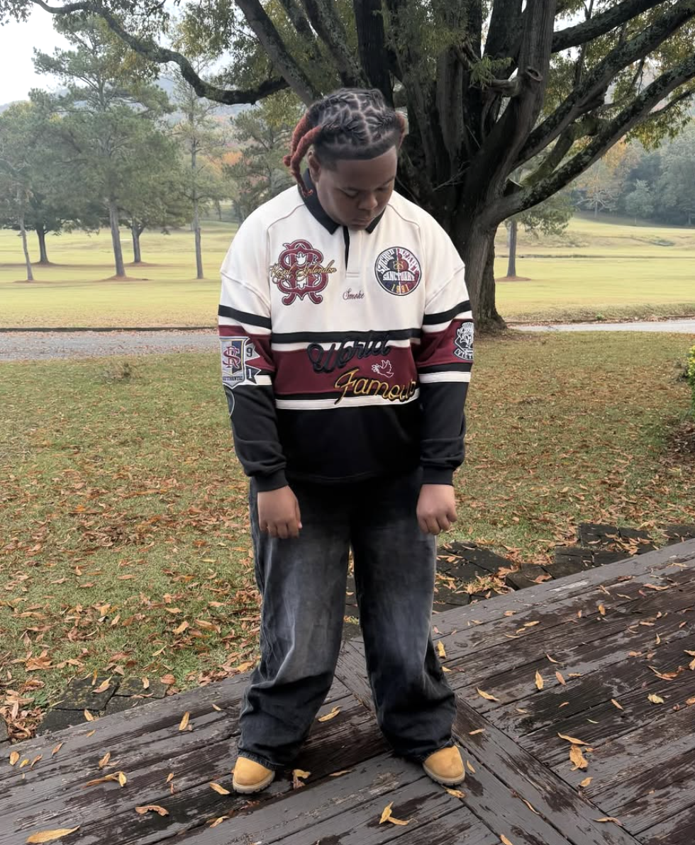 Young person with dyed hair in braids, wearing a colorful varsity jacket, looking down, standing on a wooden deck outdoors with fallen leaves, grass, trees, and a park in the background.