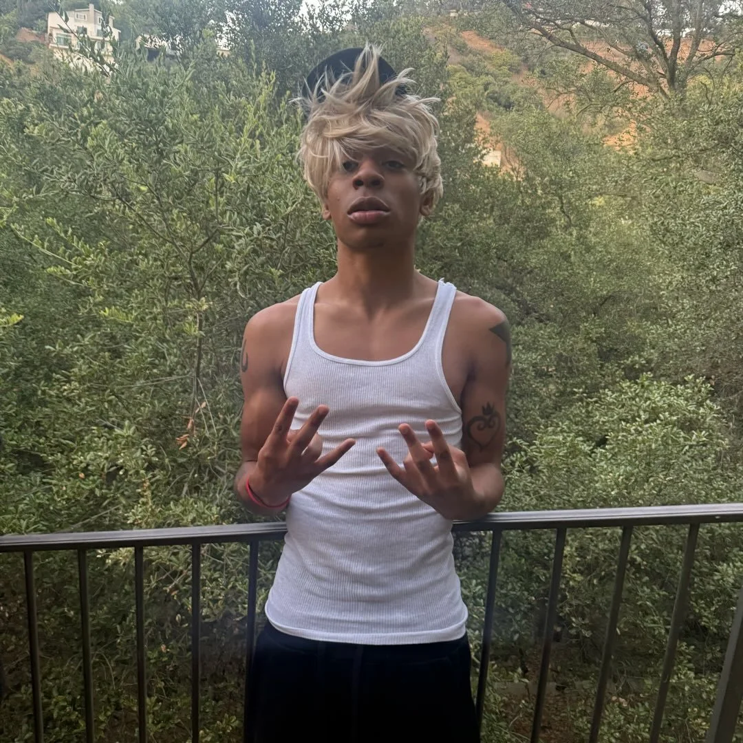 Young man with long blond hair wearing a white tank top and black hat, standing outdoors on a balcony with green trees in the background, making a hand gesture.