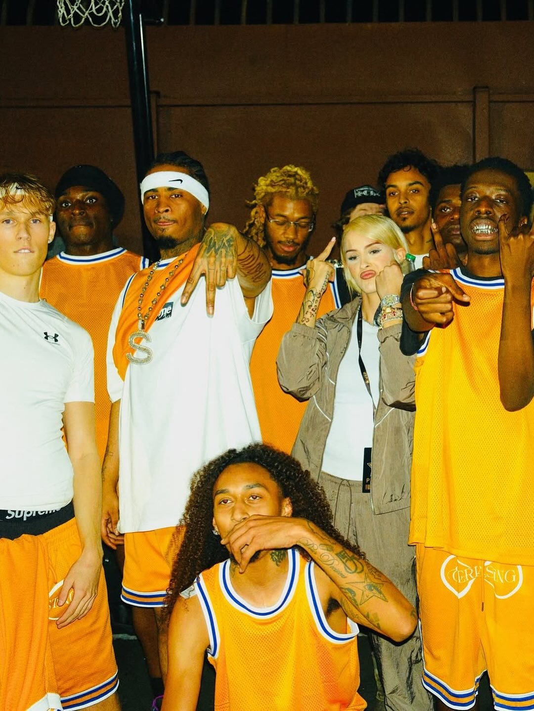 Group of rappers at the Underground Sound & Hyperpop Daily Basketball Tournament, in basketball uniforms, posing together with gestures, in a gymnasium or indoor sports arena.