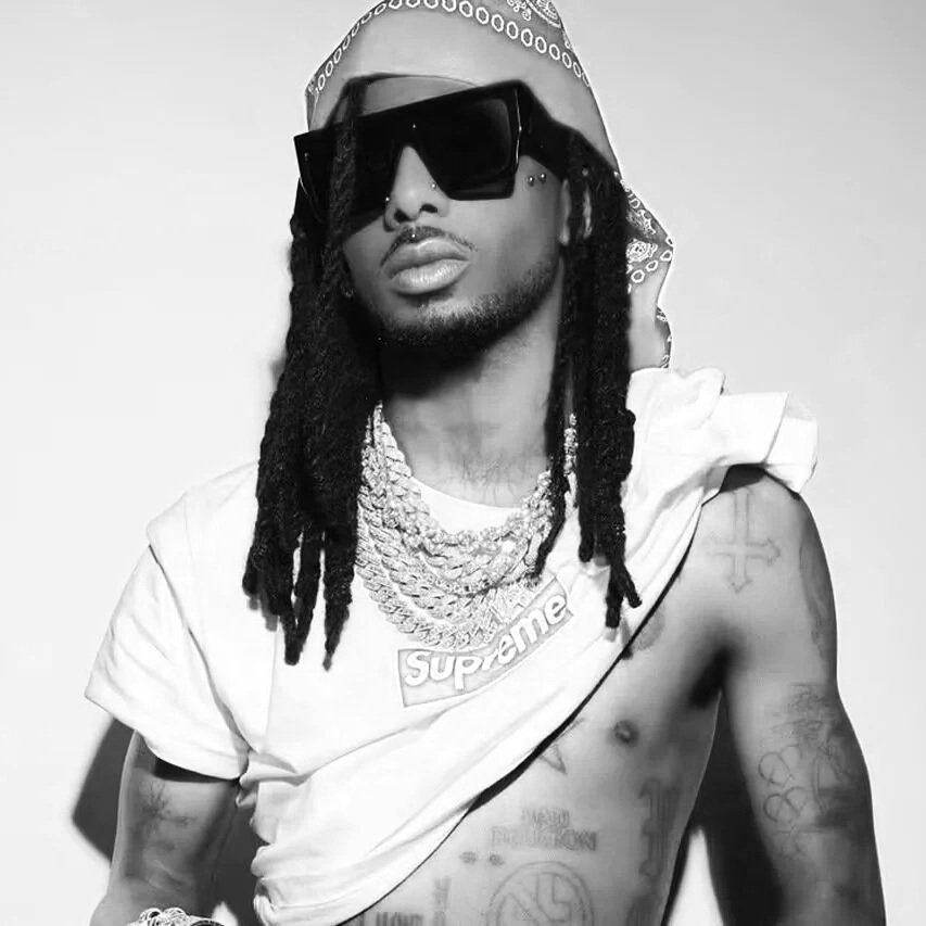 A man with dreadlocks wearing large sunglasses, jewelry, a cap, and a casual sleeveless shirt with a Supreme logo, showing tattoos on his chest and arms.